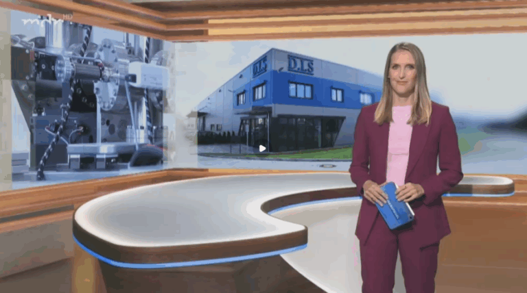 Frau mit blondem Haar in lila Anzug lächelt vor Studiobühne mit Bild von Firmengebäude.