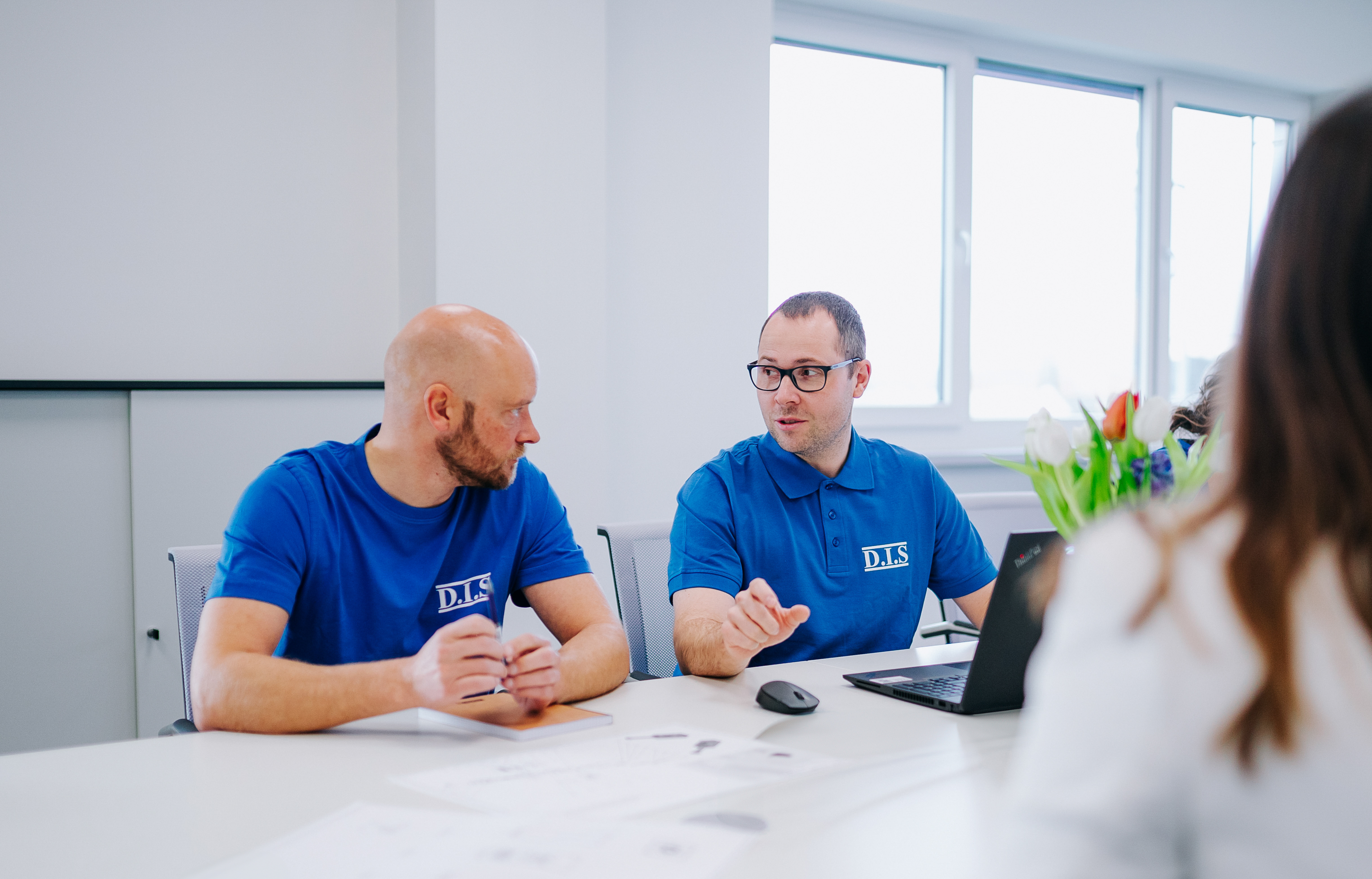 Zwei Männer in blauen Poloshirts, die an einem Tisch diskutieren.