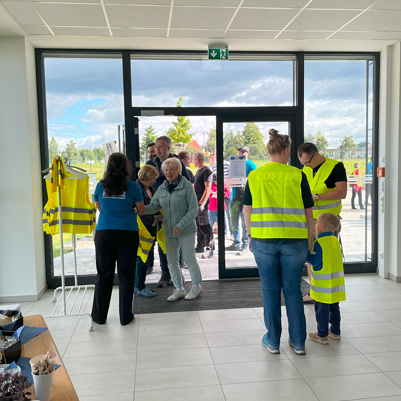 Eine Gruppe von Menschen in leuchtend gelben Westen steht lächelnd in einem modernen Eingangsbereich mit großem Fenster.