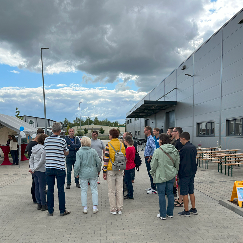 Eine Gruppe von Personen steht im Freien, diskutiert entspannt vor einem Gebäude unter bewölktem Himmel.