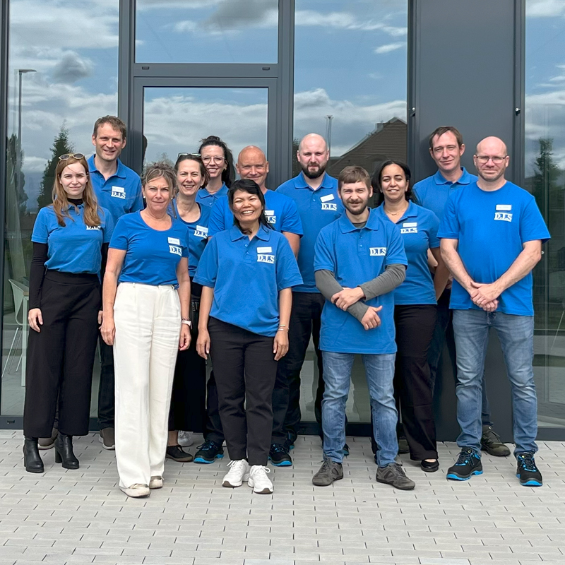 Eine Gruppe von elf lächelnden Menschen in blauen Hemden vor einer Glasfassade