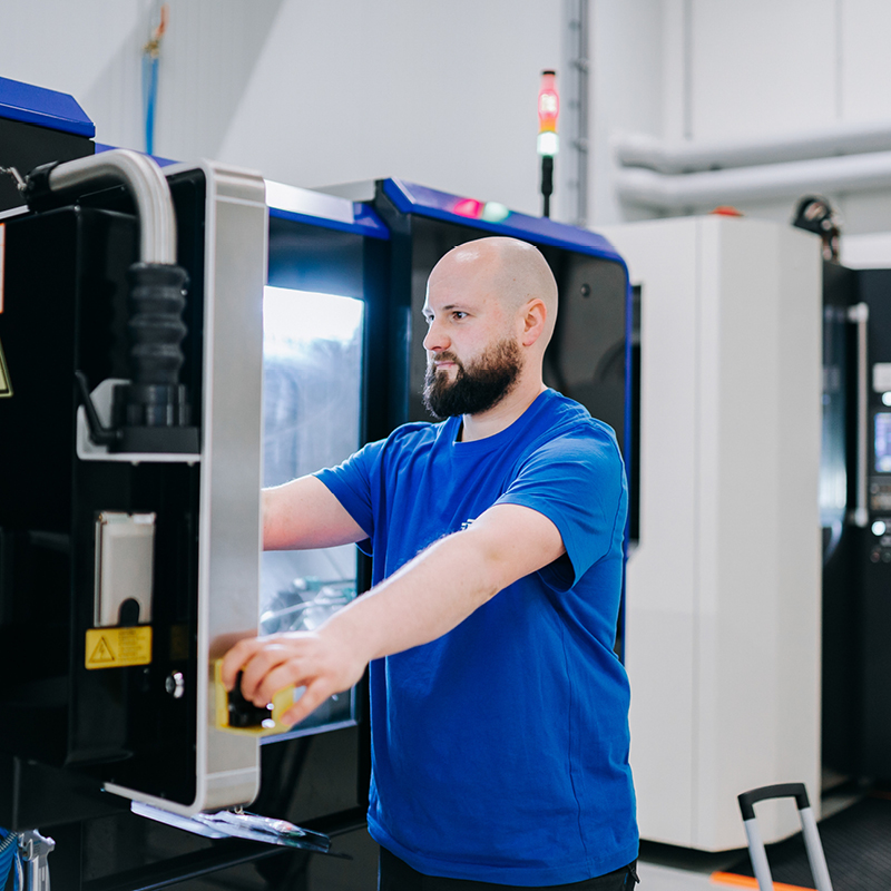Ein bärtiger Mann mit ernster Miene bedient eine CNC Maschine im leuchtend blauen Shirt